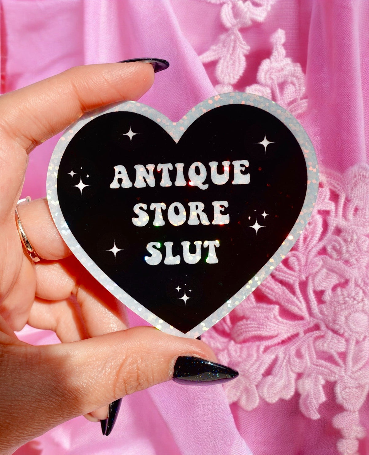 Funny Heart sticker held in hand, black with silver border; 'ANTIQUE STORE SLUT' lettering