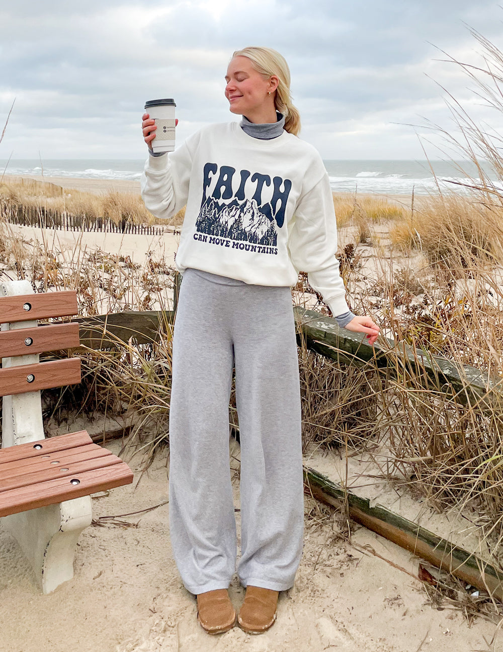Woman wearing a Christian Crewneck Sweatshirt with a faith design, enjoying a coffee on the beach
