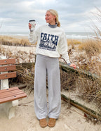 Woman wearing a Christian Crewneck Sweatshirt with a faith design, enjoying a coffee on the beach