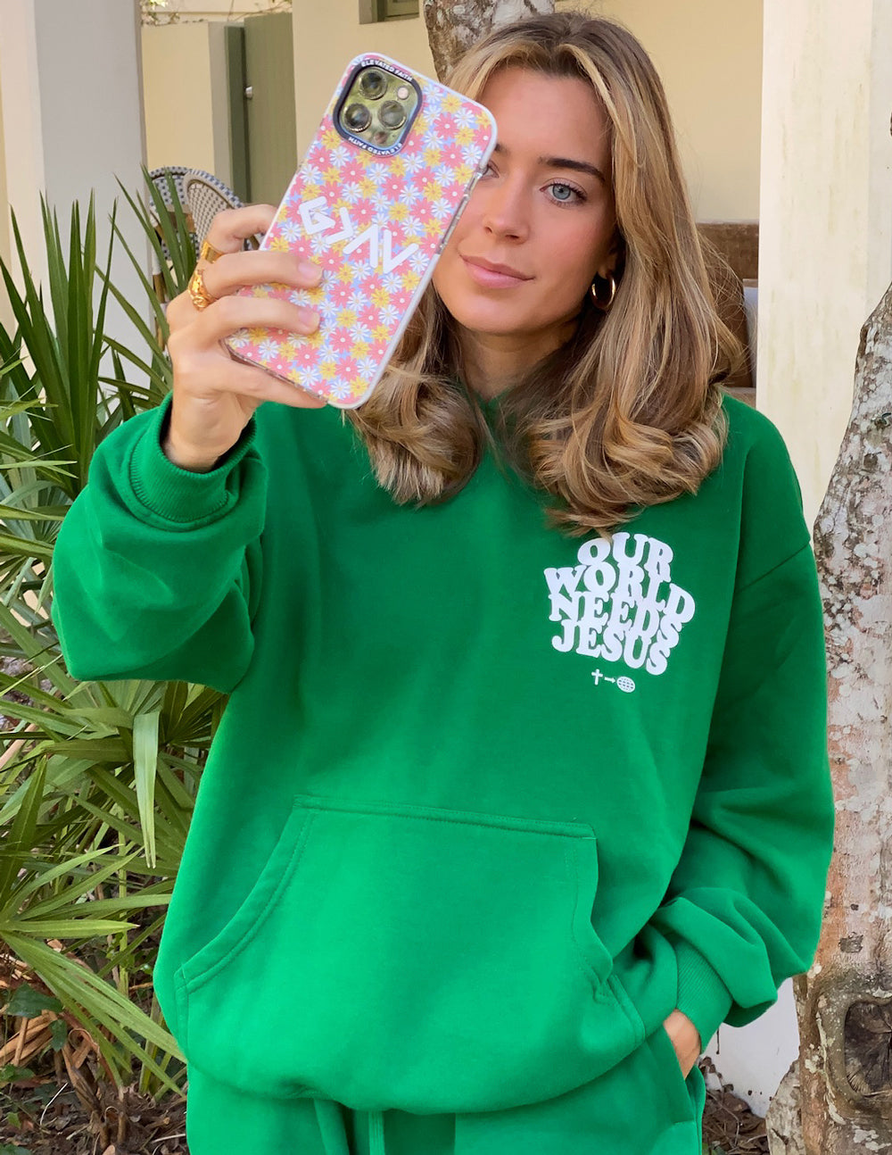 A woman holding a colorful Christian iPhone Case with a floral design while wearing a green hoodie