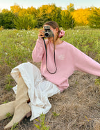 Woman wearing a pink Christian Crewneck Sweatshirt in a grassy field, holding a vintage camera