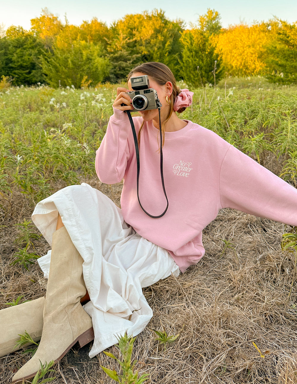 Woman wearing a pink Christian Crewneck Sweatshirt in a grassy field, holding a vintage camera