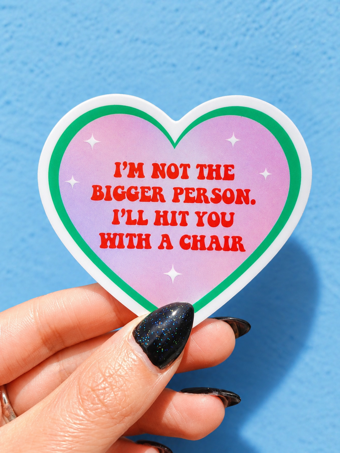 Funny Heart-shaped sticker with pink design, green border, red quote text, held in fingers