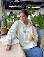 Woman in a cream Christian Hoodie with Freedom print, enjoying coffee in a cozy café