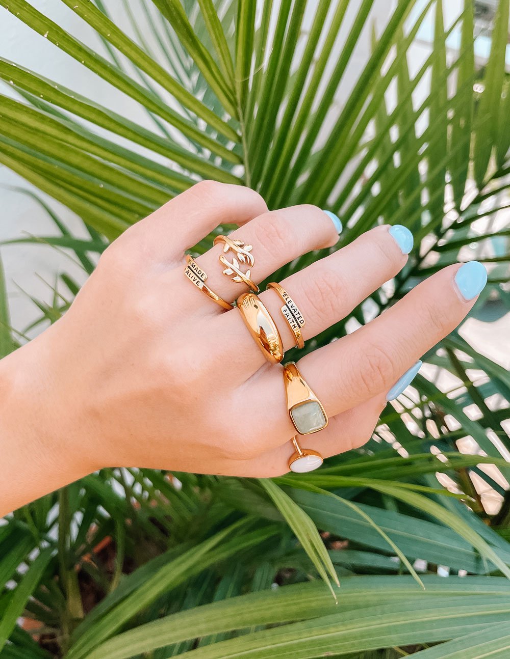 Elevated Faith Christian Jewelry featuring stylish gold rings displayed on a hand among tropical leaves