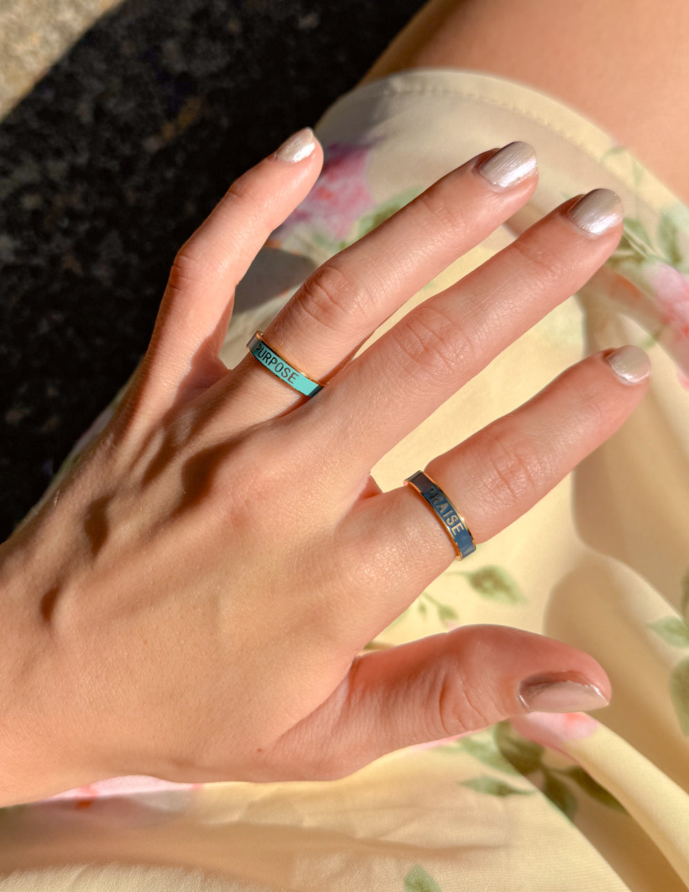 Elevated Faith Christian Jewelry Rings in Blue and Gold on a Hand Against Floral Background
