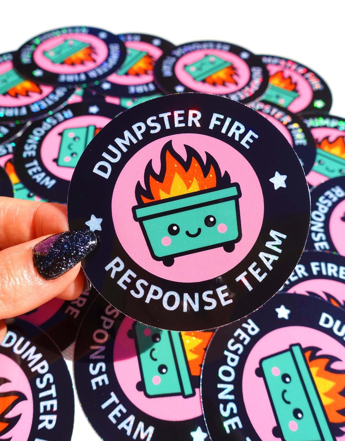 Funny Hand holding round black sticker with pink fire pot badge and “DUMPSTER FIRE RESPONSE TEAM”