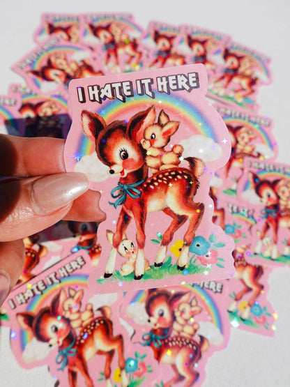 Funny Cartoon fox 'I HATE IT HERE' sticker with rainbow and clouds, held in hand