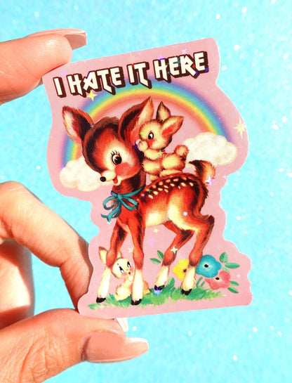 Funny Cartoon foxes 'I HATE IT HERE' sticker with rainbow and clouds, held between fingers