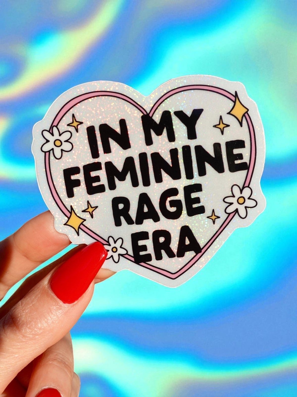 Funny Heart-shaped glitter sticker: 'In My Feminine Rage Era', held by red-nailed hand