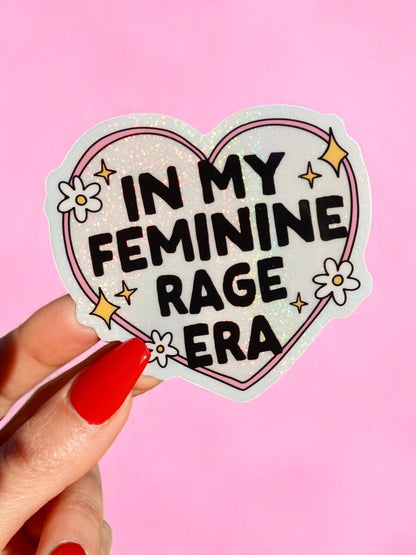 Funny Heart-shaped glitter sticker with 'In My Feminine Rage Era' text, held by hand