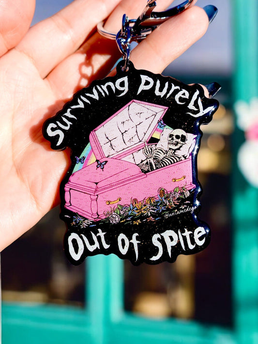 Funny Black and pink coffin-themed keychain with skull and text “Surviving Purely Out of Spite”