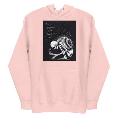 Light pink pullover hoodie with the 'Tiny Moments' goth graphic: a kneeling skeleton holding a tiny pink flower on a black background, with the words "Tiny moments hold the loudest magic"