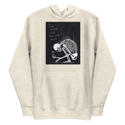 Oatmeal heather pullover hoodie with the 'Tiny Moments' goth graphic: a kneeling skeleton holding a tiny pink flower on a black background, with the words "Tiny moments hold the loudest magic"