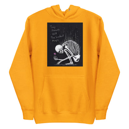Gold pullover hoodie with the 'Tiny Moments' goth graphic: a kneeling skeleton holding a tiny pink flower on a black background, with the words "Tiny moments hold the loudest magic"