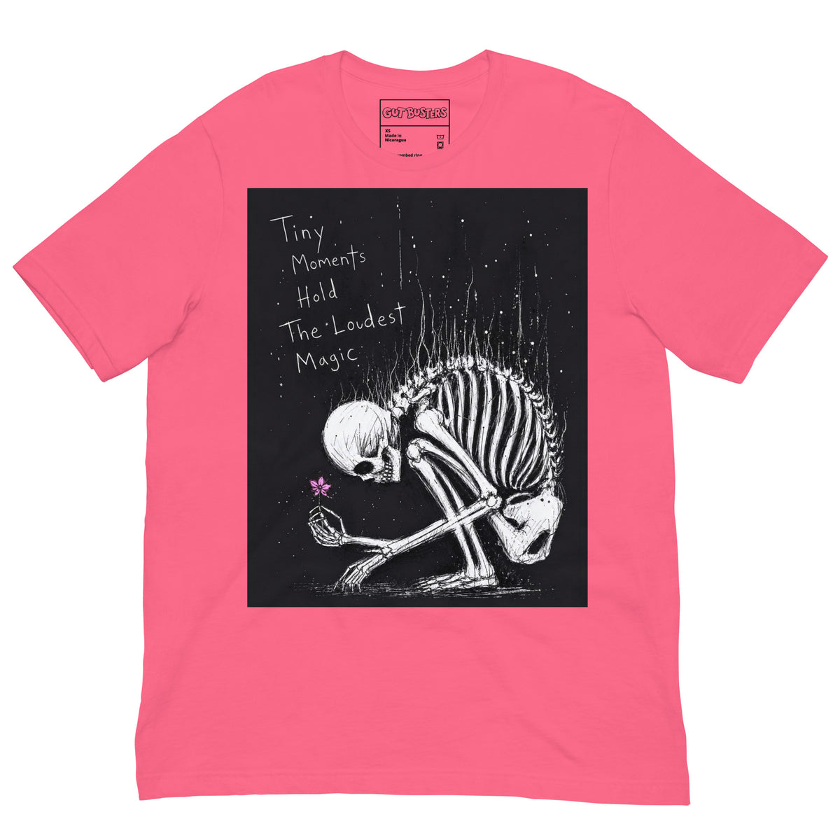 Pink unisex t-shirt with the 'Tiny Moments' goth graphic: a kneeling skeleton holding a tiny pink flower on a black background, with the words "Tiny moments hold the loudest magic"
