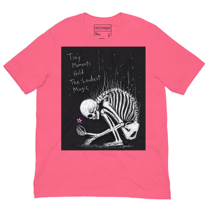 Pink unisex t-shirt with the 'Tiny Moments' goth graphic: a kneeling skeleton holding a tiny pink flower on a black background, with the words "Tiny moments hold the loudest magic"