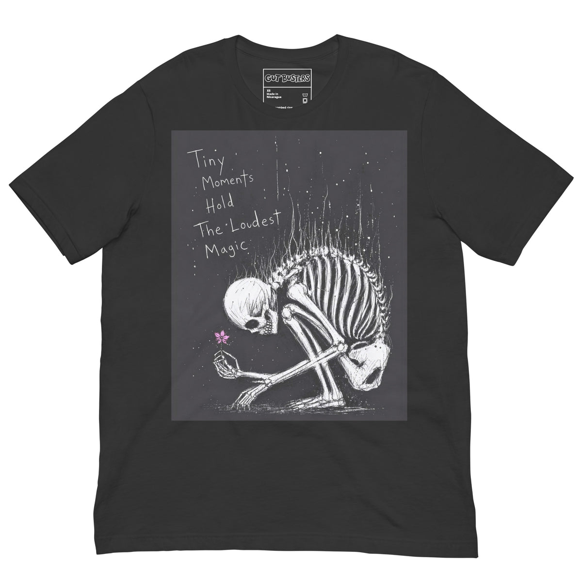Dark grey unisex t-shirt with the 'Tiny Moments' goth graphic: a kneeling skeleton holding a tiny pink flower on a black background, with the words "Tiny moments hold the loudest magic"