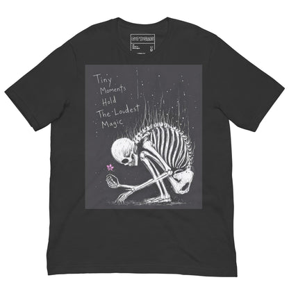 Dark grey unisex t-shirt with the 'Tiny Moments' goth graphic: a kneeling skeleton holding a tiny pink flower on a black background, with the words "Tiny moments hold the loudest magic"