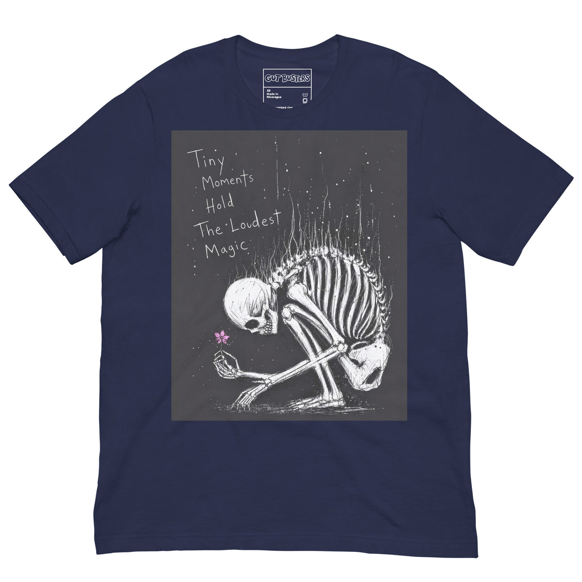Navy unisex t-shirt with the 'Tiny Moments' goth graphic: a kneeling skeleton holding a tiny pink flower on a black background, with the words "Tiny moments hold the loudest magic"