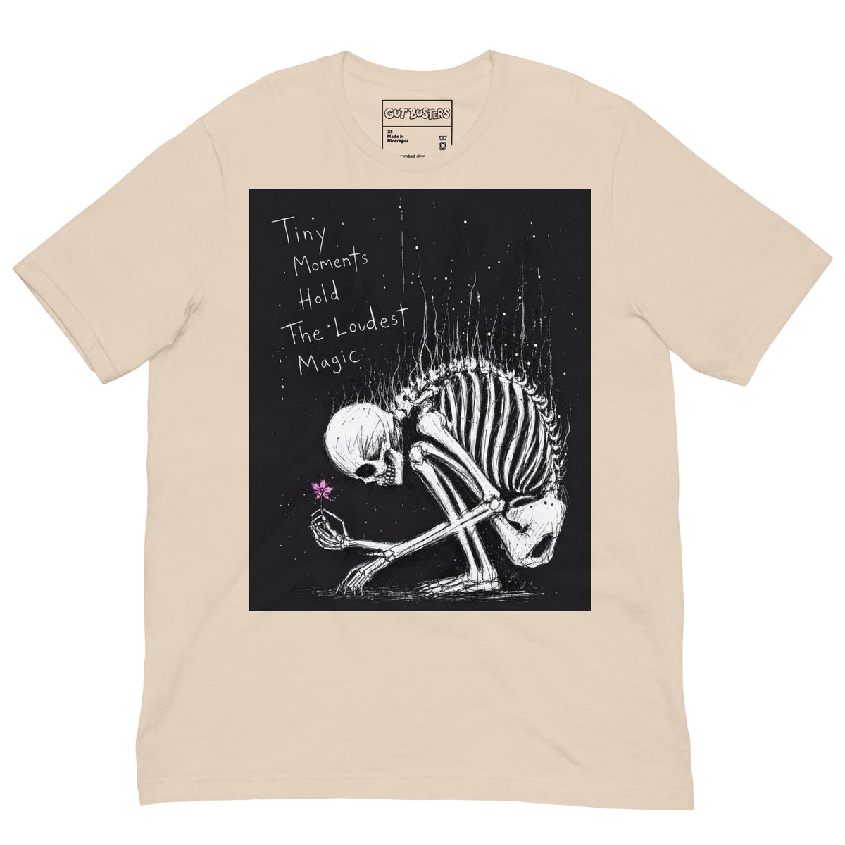 Soft cream unisex t-shirt with the 'Tiny Moments' goth graphic: a kneeling skeleton holding a tiny pink flower on a black background, with the words "Tiny moments hold the loudest magic"