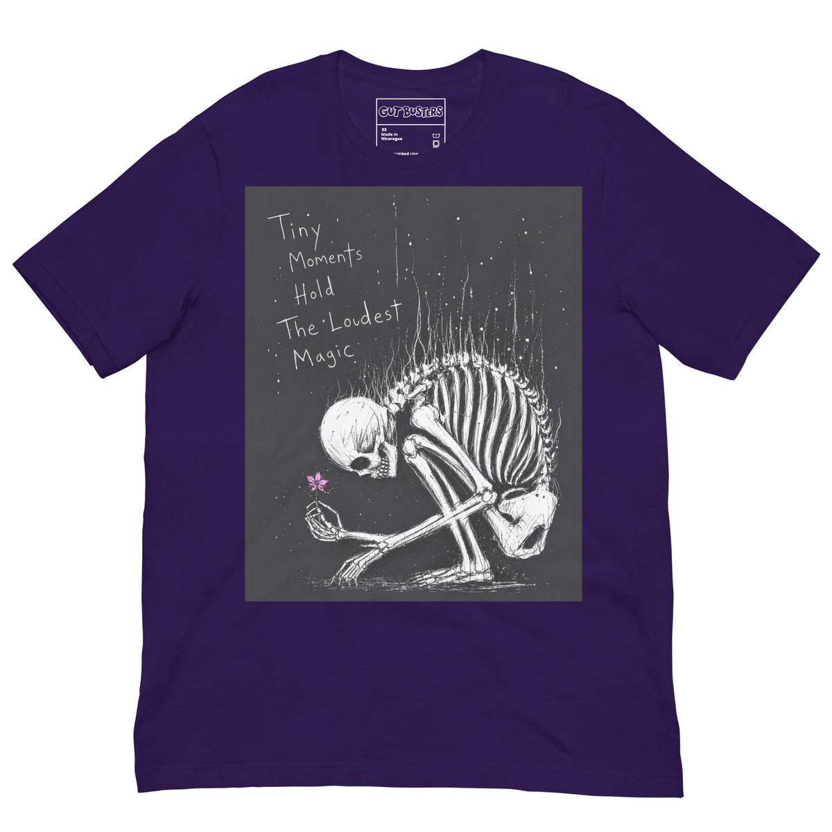 Purple unisex t-shirt with the 'Tiny Moments' goth graphic: a kneeling skeleton holding a tiny pink flower on a black background, with the words "Tiny moments hold the loudest magic"